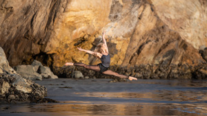 A dancer leaping on the beach in front of a large rock.