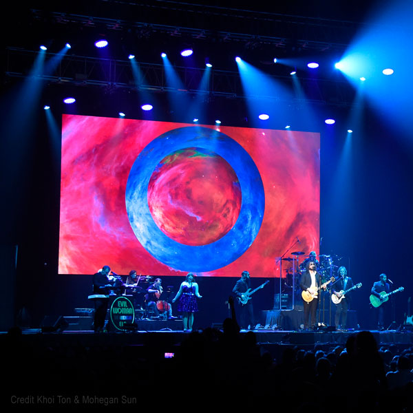 Band members of The American ELO perform in front of a giant O projection.