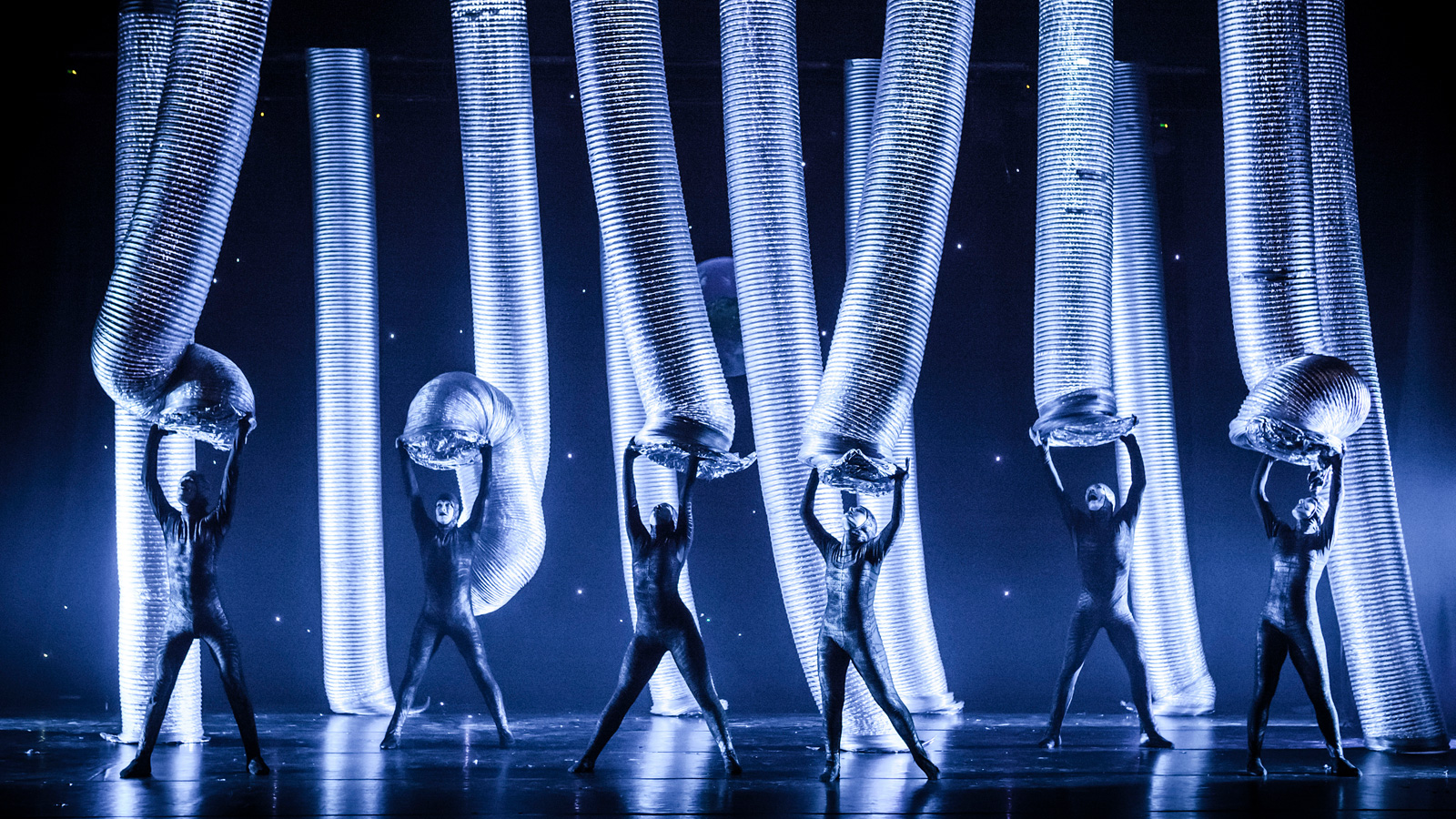 Performers dance onstage with giant aluminum tubes.