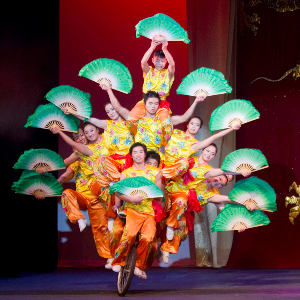 Many acrobats riding a bike while holding hand fans.