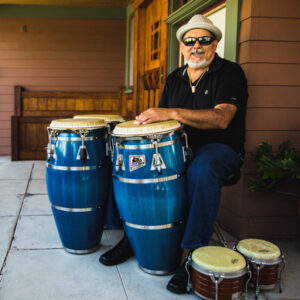 Poncho Sanchez seated on a porch with his drums.